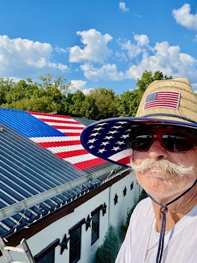 Chris Opp (man wearing a straw hat and sunglasses) with view of a mural of an U. S. A. Banner on a roof