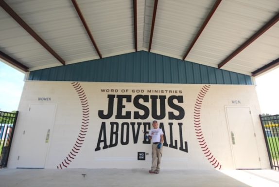 Mural with "Jesus Above All" on side of wall next to baseball park